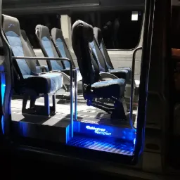 Wnętrze nowoczesnego busa z niebieskimi, pikowanymi siedzeniami i podświetlanymi schodkami wejściowymi z napisem 'Debbestrap Turistfart'.