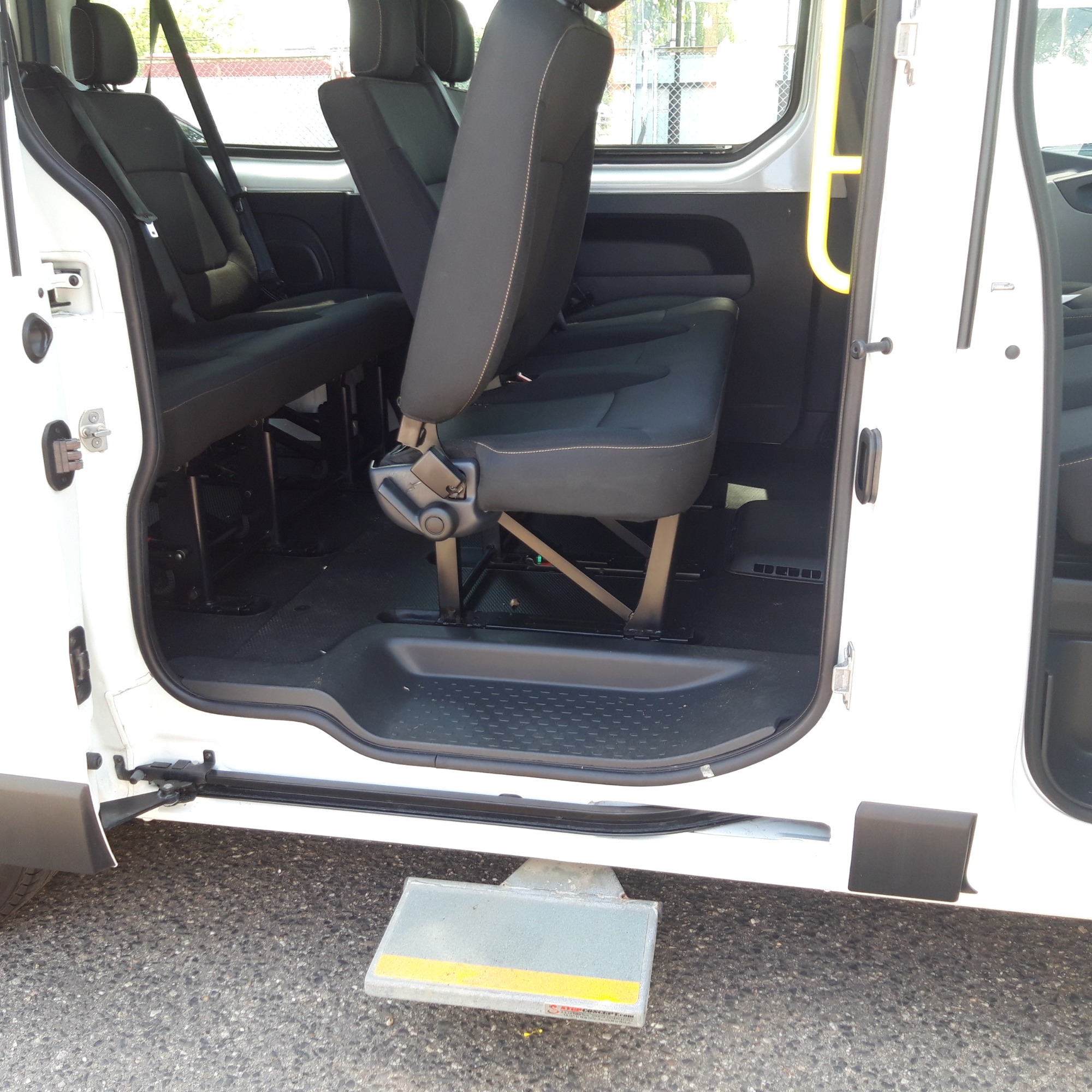 Wnętrze białego busa z widocznymi czarnymi siedzeniami z regulowanymi zagłówkami i pasami bezpieczeństwa oraz wysuwanym stopniem z żółtym paskiem ułatwiającym wsiadanie.