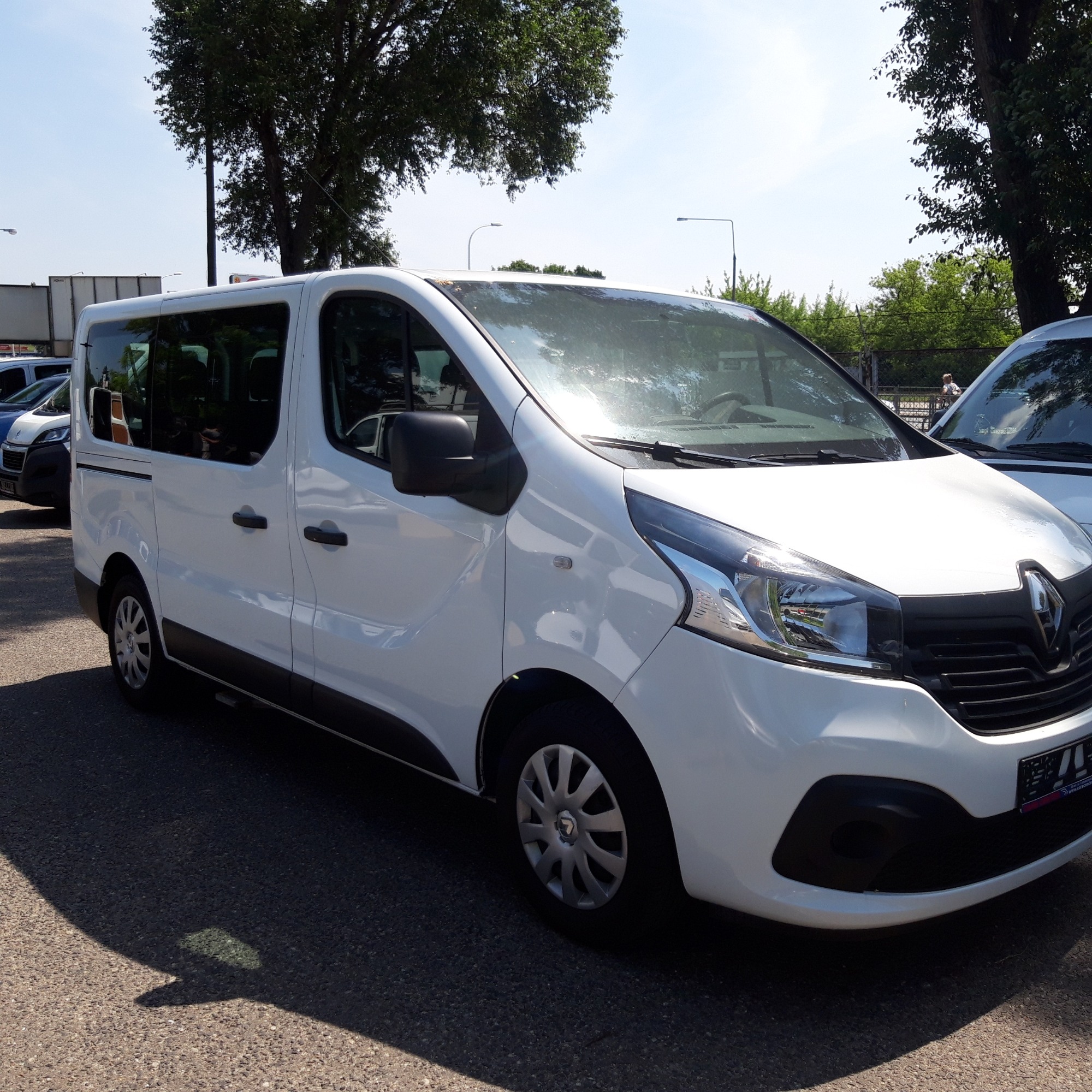 Biały Renault Trafic, zaparkowany na parkingu w słoneczny dzień, widoczny z przodu i z boku, z drzewami w tle.