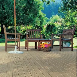 Kompozytowa deska tarasowa w odcieniach beżu, ułożona na zewnątrz, z drewnianymi krzesłami i stolikiem pod parasolem ogrodowym, na tle zieleni.