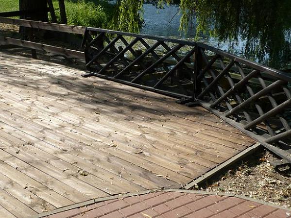 Drewniany taras z balustradą w kratkę, widok na wodę i zieleń w tle, deski tarasowe z widocznymi śladami użytkowania i drobnymi zanieczyszczeniami.