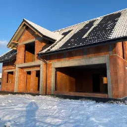 Surowy stan domu z czerwonej cegły w zimowej scenerii, częściowo pokryty śniegiem dach z czarnej dachówki, widoczne betonowe nadproża okienne i garażowe, błękitne niebo w tle.