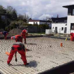 Prace zbrojarskie na przygotowanym fundamencie pod budynek, widoczni pracownicy w czerwonych kombinezonach układający siatkę zbrojeniową, w tle domy jednorodzinne i częściowo zachmurzone niebo.