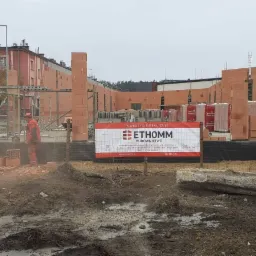 Wznoszenie ścian z czerwonej cegły Porotherm na placu budowy. Widoczny pracownik w czerwonym kombinezonie, rusztowania, palety z cegłami oraz tablica informacyjna firmy budowlanej Ethomm.