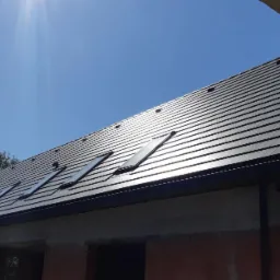 Czarny dach z dachówkami i oknami dachowymi na budynku w trakcie budowy, widok z dołu pod słońce.