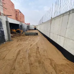 Wykop z betonowymi ścianami i fundamentami w trakcie budowy, widoczny częściowo w oddali mały żółty sprzęt budowlany i elementy zbrojeniowe.