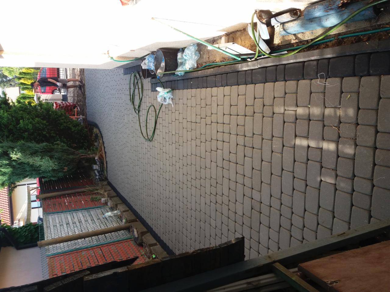 Ułożona szara kostka brukowa z czarnym obrzeżem, widoczne węże ogrodowe, wiadra i worki z cementem przy ścianie budynku w trakcie prac wykończeniowych.