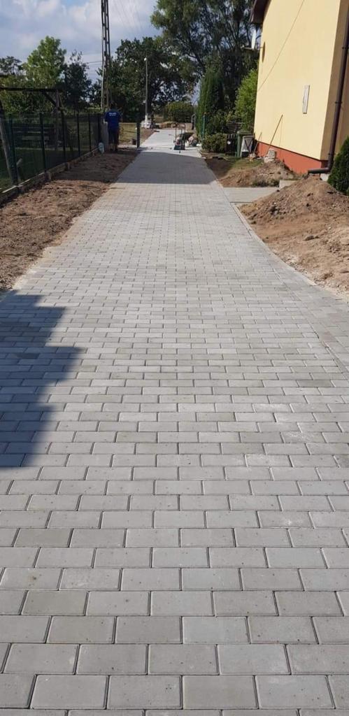 Długa, prosta ścieżka wyłożona szarą kostką brukową prowadząca wzdłuż domu z widocznymi kupkami ziemi po bokach i pracownikiem w oddali.