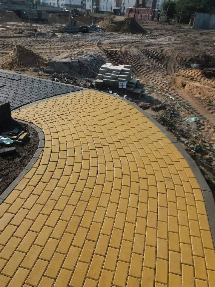 Żółta kostka brukowa ułożona łukiem, obok czarna kostka, w tle teren budowy z ziemią i stertą kostki.