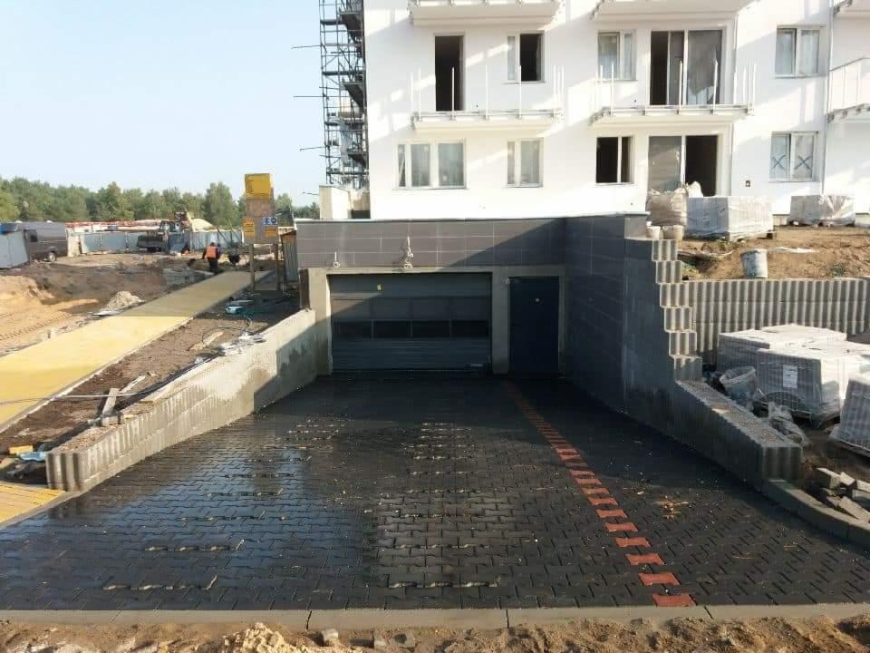 Nowa, mokra, czarna kostka brukowa prowadząca do garażu w budynku mieszkalnym, widoczna czerwona linia z kostki, palety z kostką obok, w tle plac budowy i rusztowania.