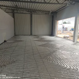 Układanie rur ogrzewania podłogowego na izolacji w surowym stanie budynku z widoczną konstrukcją stalową dachu i ścianami z bloczków.