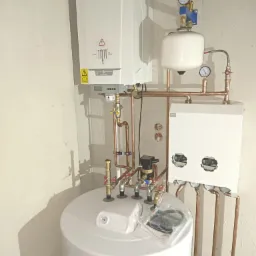 Biały wiszący piec gazowy De Dietrich z podłączonymi miedzianymi rurami, zbiornik ciśnieniowy z manometrami, biały zasobnik ciepłej wody i elementy instalacji grzewczej w kotłowni, widoczne zawory...