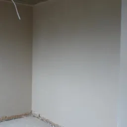 Surowe wnętrze w trakcie remontu: ściany pokryte świeżą warstwą gładzi, widoczne niedoskonałości przy podłodze, sufit z drewnianych desek, wystający kabel elektryczny.