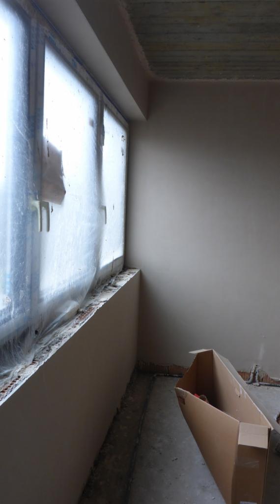 Przygotowanie do prac wykończeniowych: okno zabezpieczone folią, świeżo otynkowana ściana w odcieniach beżu, karton na podłodze.