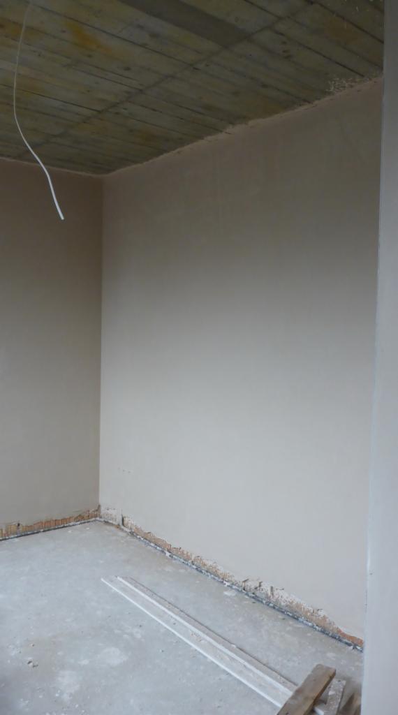 Surowe wnętrze w trakcie remontu: ściany pokryte świeżą warstwą gładzi, widoczne niedoskonałości przy podłodze, sufit z drewnianych desek, wystający kabel elektryczny.