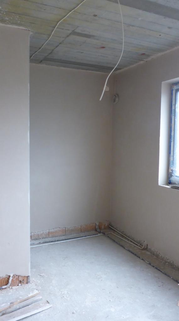 Pokój w trakcie remontu: ściany pokryte świeżym tynkiem, widoczne przewody elektryczne zwisające z sufitu, okno po prawej stronie, fragment muru pod ścianą.
