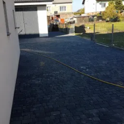 Ciemna kostka brukowa ułożona na podjeździe przed garażem, z widoczną studzienką kanalizacyjną i kartonem po soku przy krawędzi.