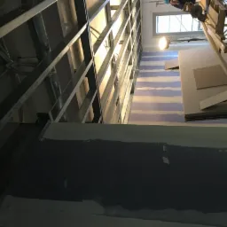 Stelaż metalowy pod konstrukcję z płyt karton-gips na tle ściany z widocznymi pasami kleju i ułożonymi płytami, widok z dołu, narzędzia pracy w tle.