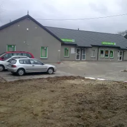 Szary budynek apteki w Lipinach z zielonymi obramowaniami okien, widoczne dwa samochody osobowe na parkingu oraz fragment terenu z ziemią.