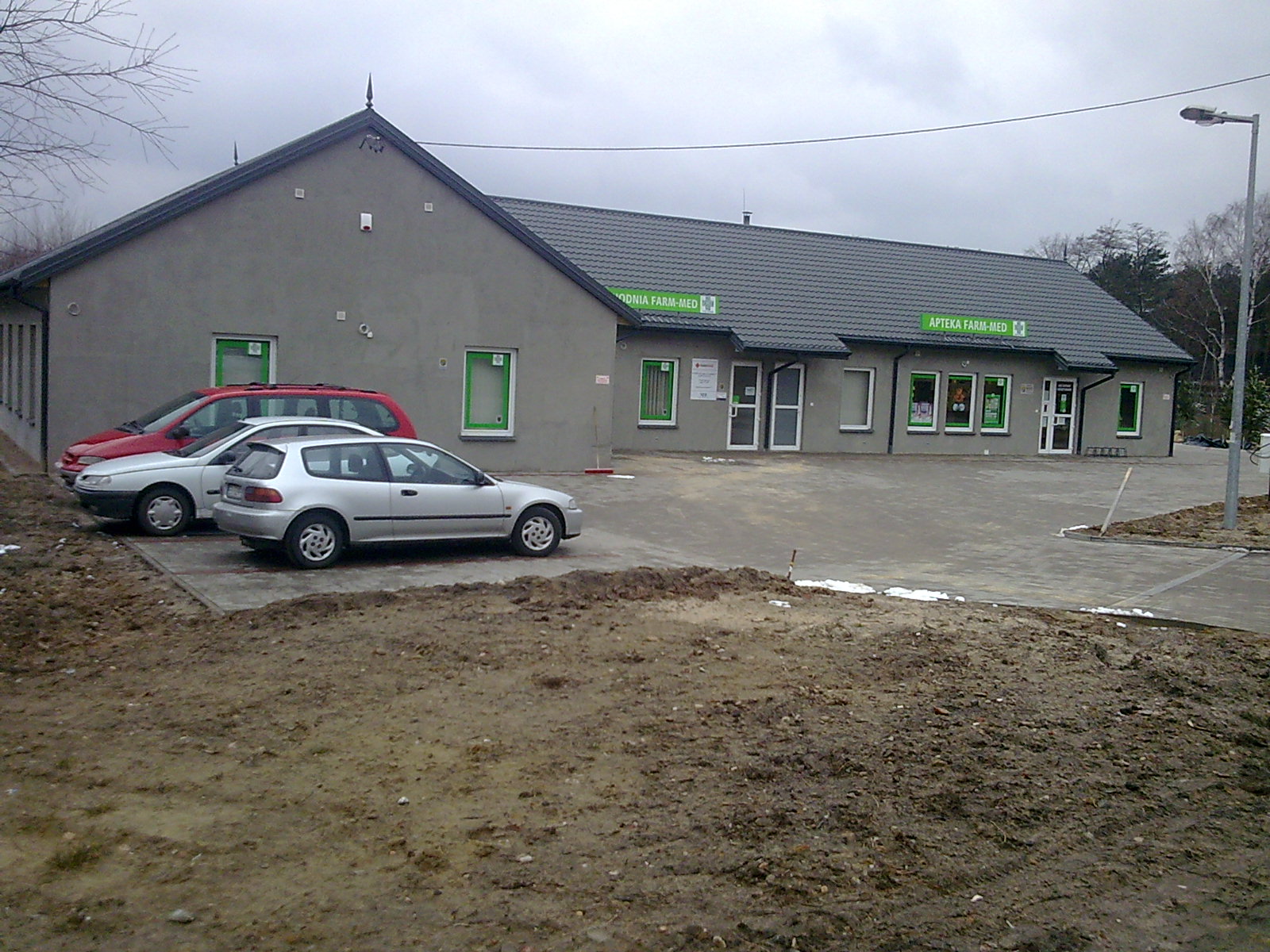 Szary budynek apteki w Lipinach z zielonymi obramowaniami okien, widoczne dwa samochody osobowe na parkingu oraz fragment terenu z ziemią.