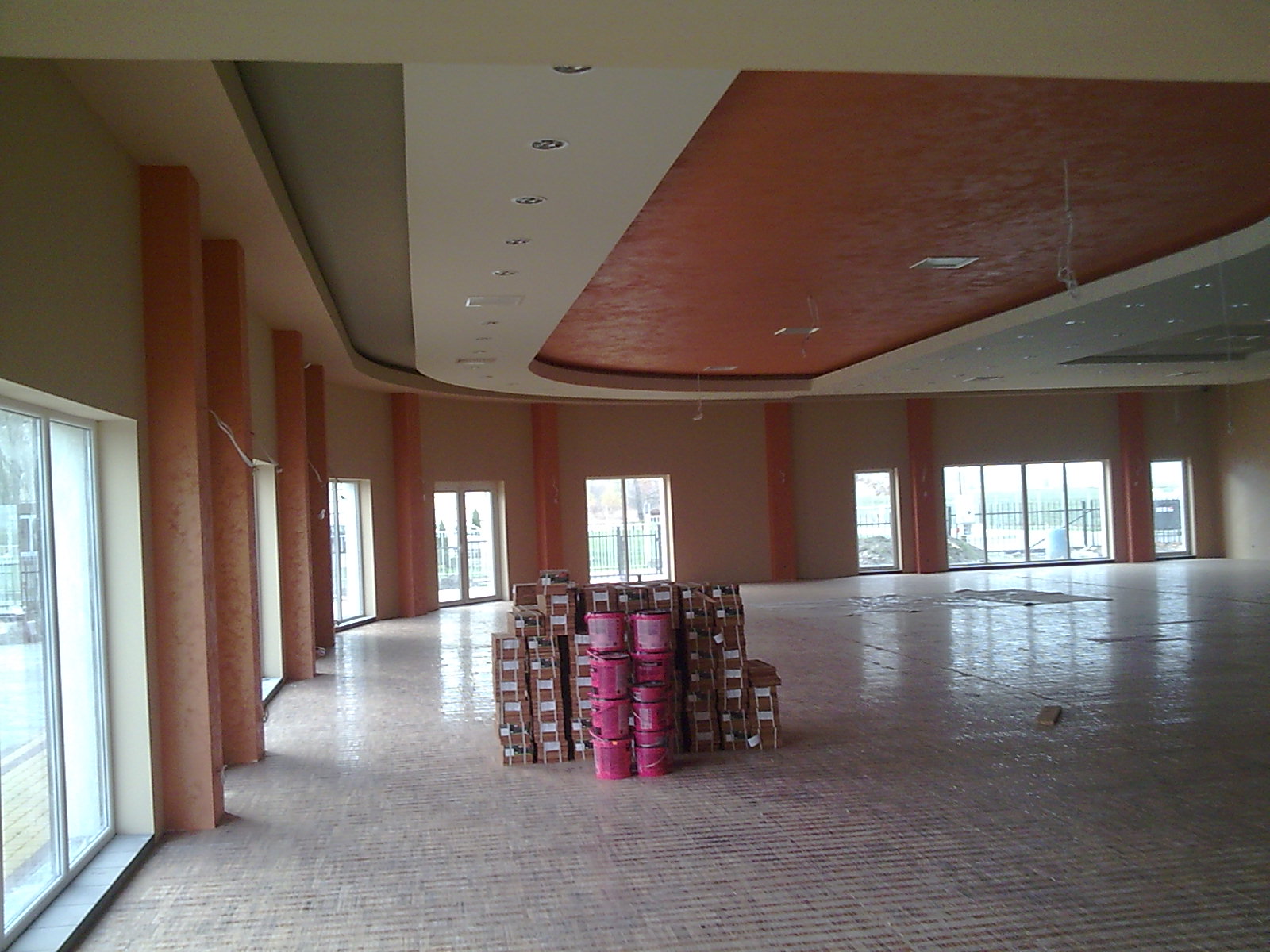 Duże, puste pomieszczenie z kolumnami w trakcie prac wykończeniowych, widoczne puszki z farbą w kolorze różowym i kartony, sufit z dekoracyjnym malowaniem w odcieniach brązu i beżu.