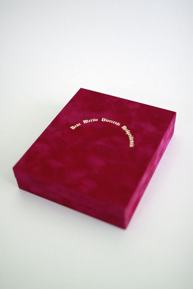 Kwadratowe, bordowe pudełko z aksamitu z napisem w kolorze złotym 'Bene Merito Dioecesis Bydgostiensis' na białym tle