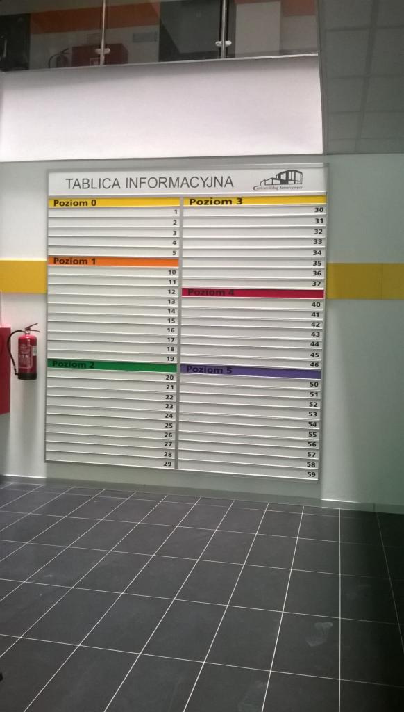 Tablica informacyjna z listą numerów poziomów i numerów miejsc w budynku, widoczna z perspektywy posadzki z ciemnych płytek.