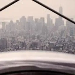 Panorama miasta z wysokości, widoczna przez metalową konstrukcję zabezpieczającą, przypominająca kratownicę. W tle liczne wieżowce spowite lekką mgłą.