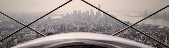 Panorama miasta z wysokości, widoczna przez metalową konstrukcję zabezpieczającą, przypominająca kratownicę. W tle liczne wieżowce spowite lekką mgłą.
