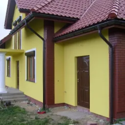 Żółty dom jednorodzinny z czerwoną dachówką i ceglanymi elementami dekoracyjnymi, widok z boku, częściowo zagospodarowany teren wokół budynku.