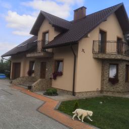 Agamedes Krzysztof Czepiel - Dwupiętrowy dom z beżową elewacją, brązowym dachem i kamiennymi akcentami, balkony z kutymi balustradami, numer domu 31A, jasny pies na trawniku, błękitne niebo z chmurami w tle.