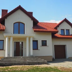 Jasny, dwukondygnacyjny dom jednorodzinny z czerwoną dachówką, kolumnami przy wejściu i podmurówką w odcieniach beżu.