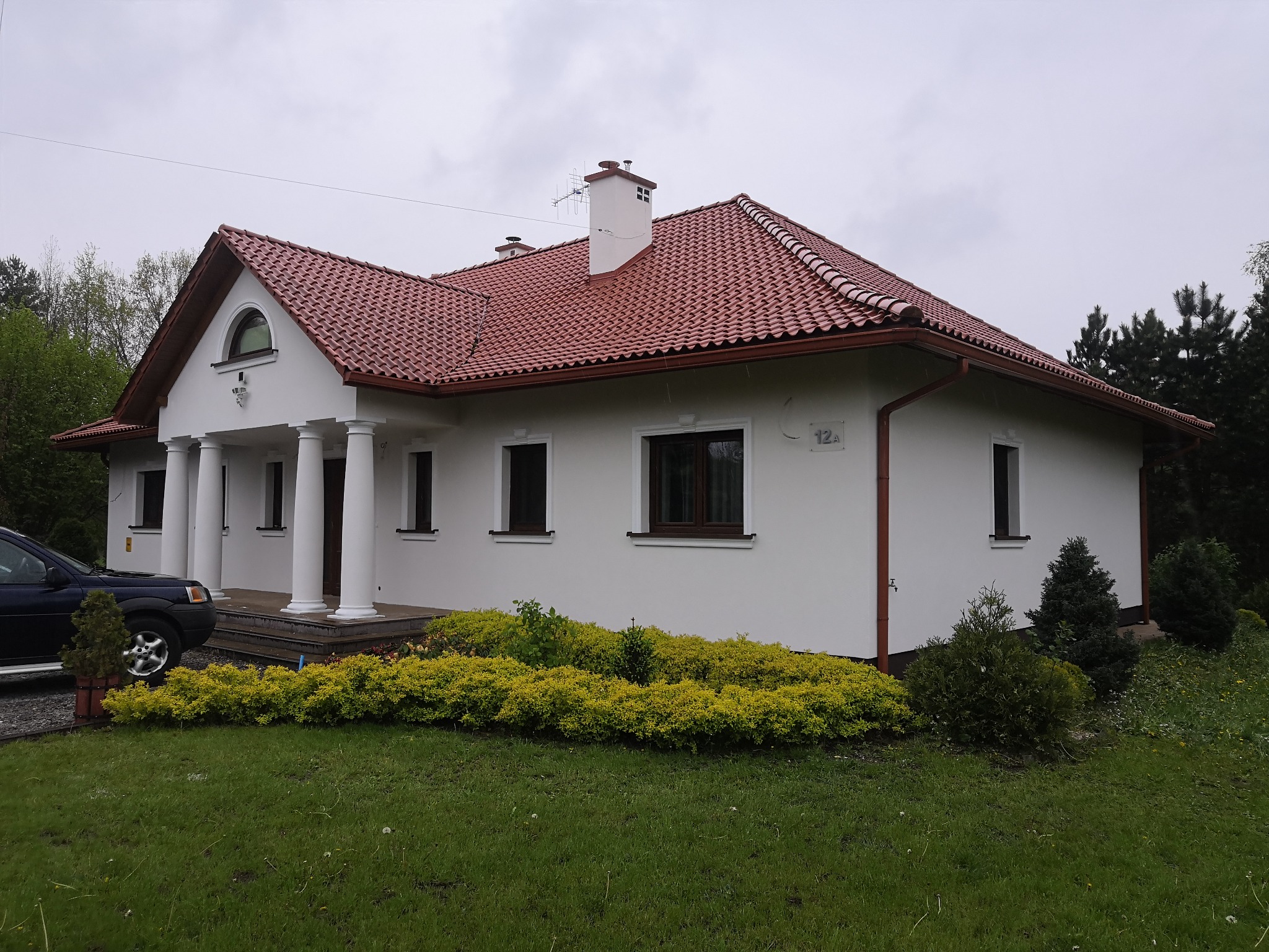 Jednopiętrowy dom z białymi ścianami, czerwoną dachówką i kolumnami przy wejściu, numer domu 12a, widoczne brązowe rynny i okna, ciemny samochód na podjeździe, zadbany trawnik i krzewy.