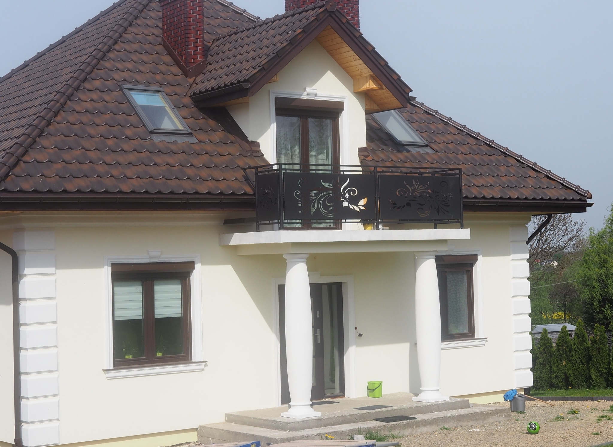 Jasna elewacja domu jednorodzinnego z brązowym dachem, balkonem z ozdobną balustradą i kolumnami, widoczne okna dachowe i ceglany komin