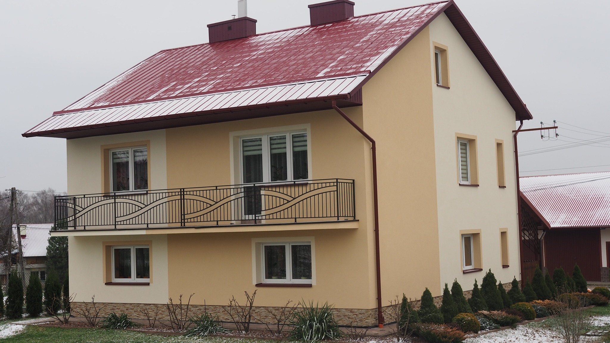 Dwukondygnacyjny dom z balkonem i czerwoną blachodachówką, elewacja w odcieniach beżu, ozdobne krzewy wzdłuż fundamentów, pochmurne niebo.