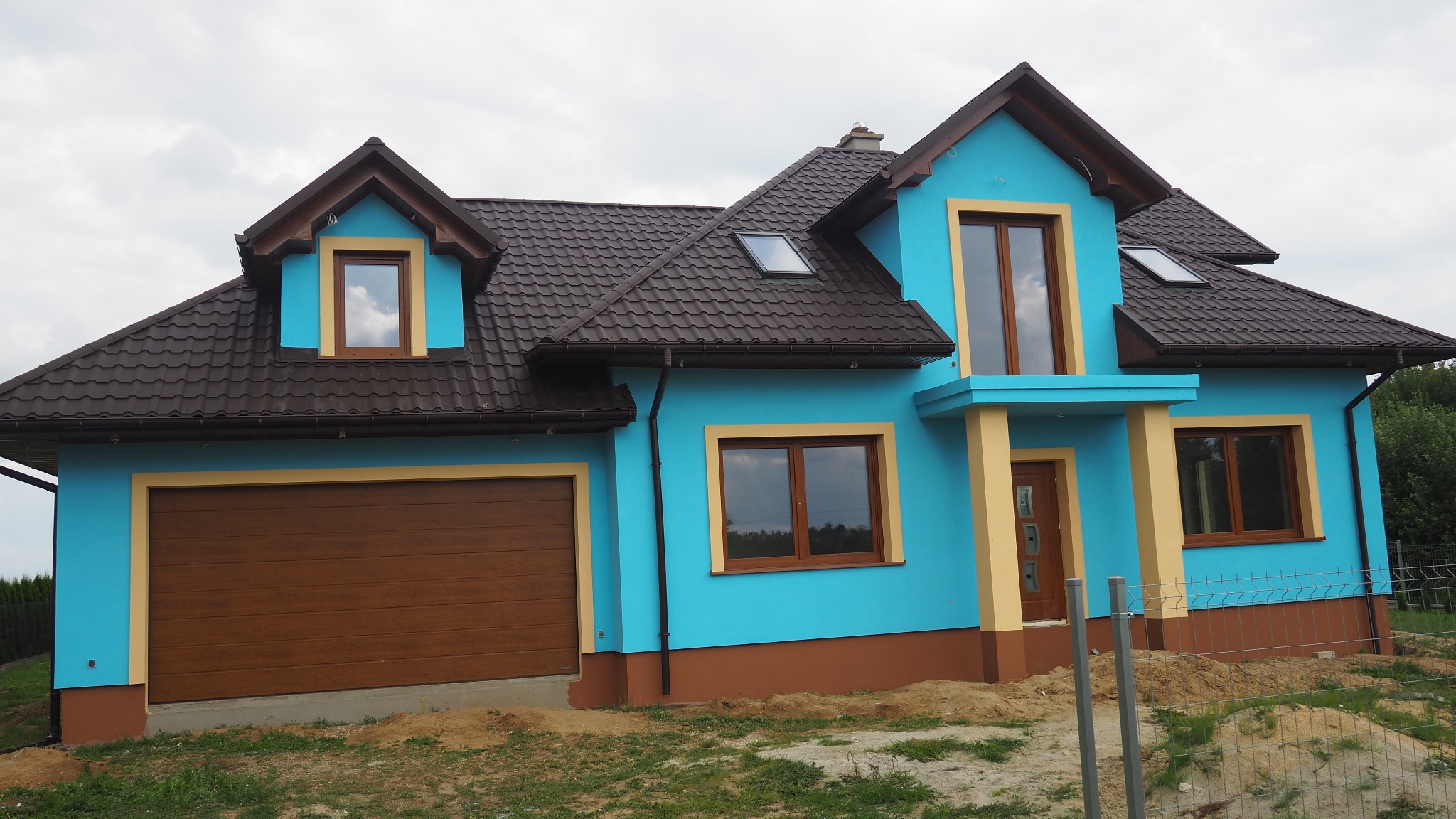 Niebieski dom jednorodzinny z brązowym dachem i garażem, żółte obramowania okien i drzwi, widok z zewnątrz na tle pochmurnego nieba.
