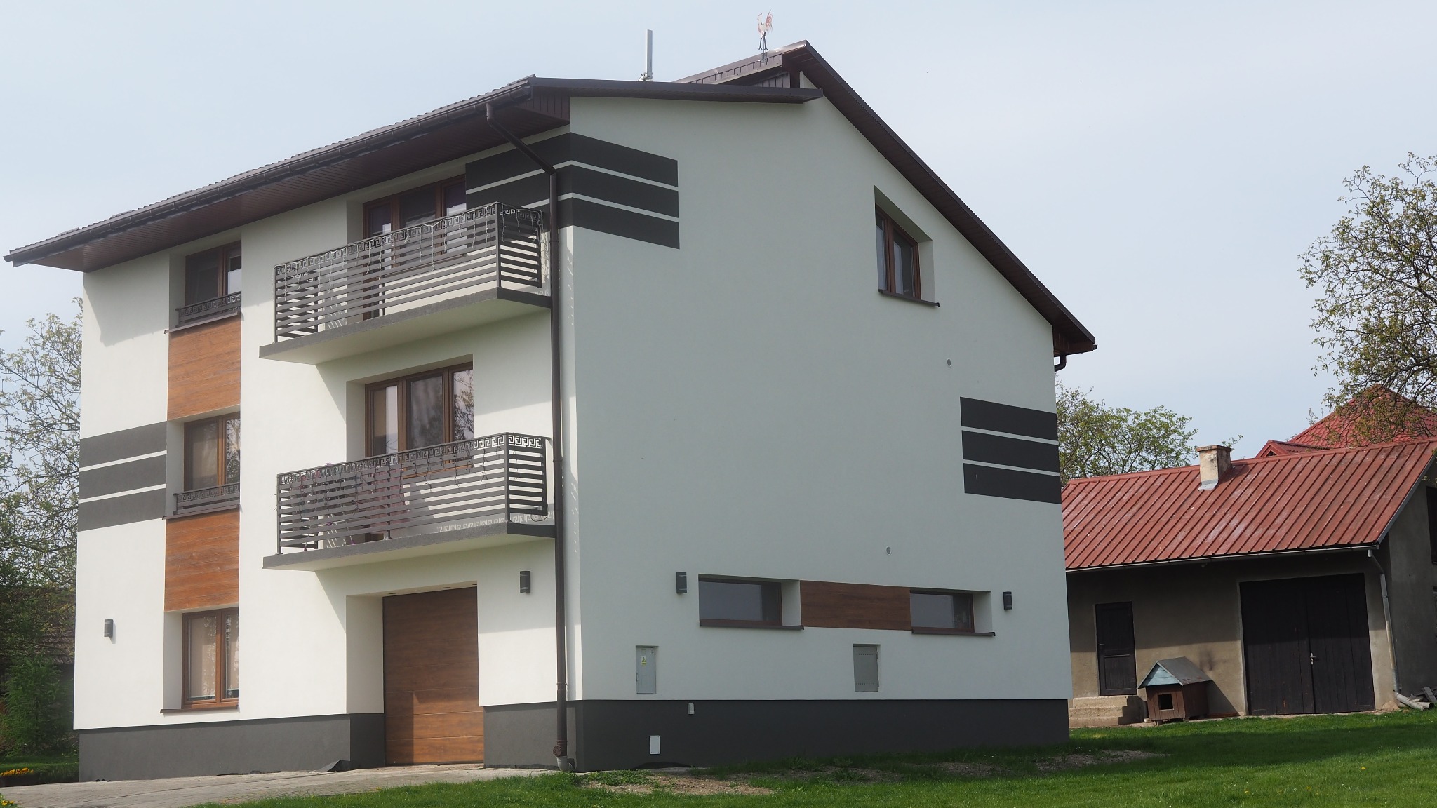 Dwupiętrowy dom z białymi ścianami, drewnianymi akcentami i ciemnymi poziomymi pasami, widoczne balkony z ozdobnymi balustradami, garaż z drewnianą bramą, w tle budynek gospodarczy z czerwonym dachem.