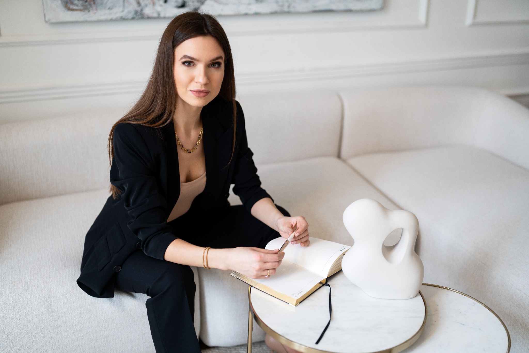 Kobieta w czarnym garniturze siedzi na kremowej sofie, przeglądając kalendarz na marmurowym stoliku z abstrakcyjną białą rzeźbą obok.