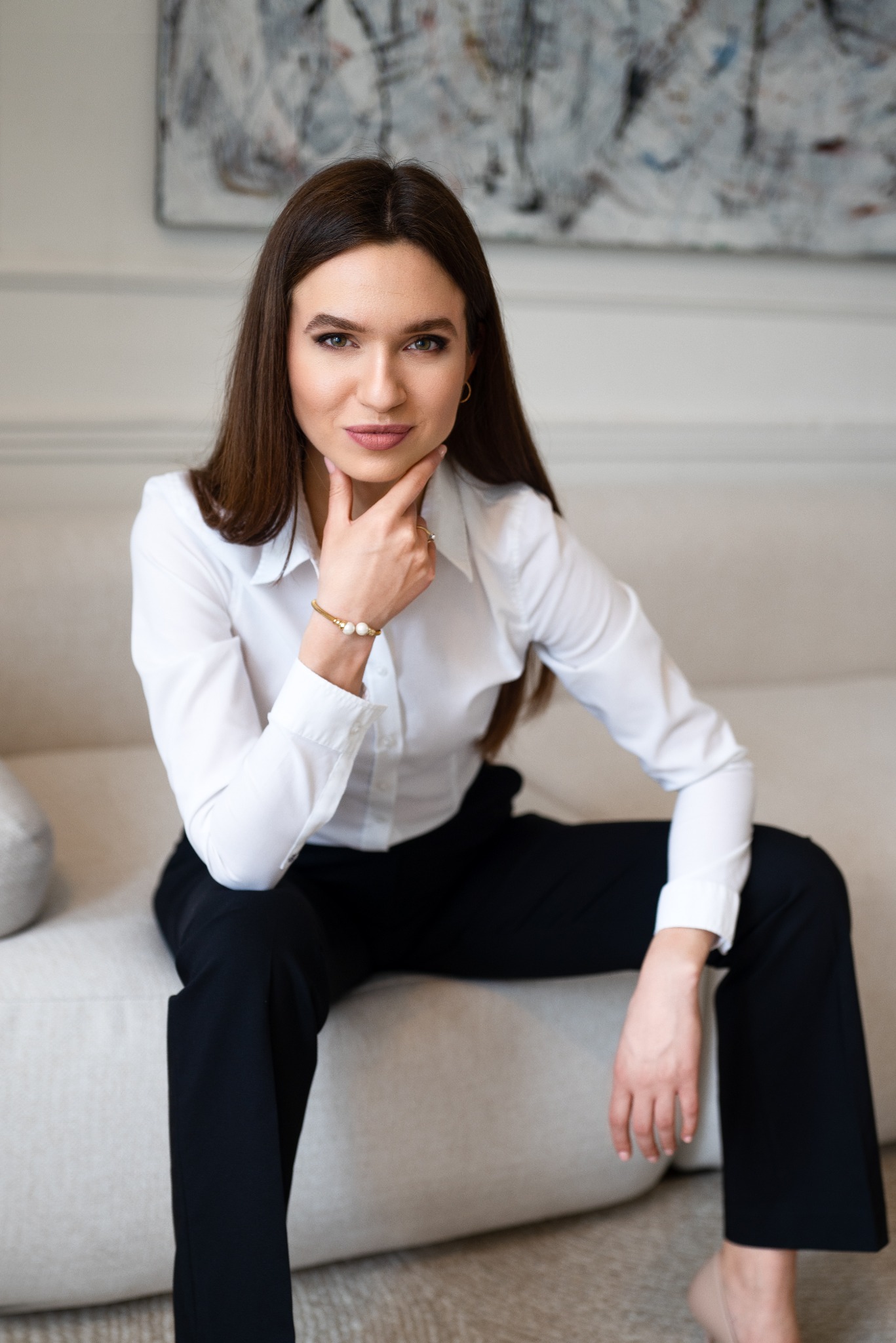Portret kobiety w białej koszuli i czarnych spodniach siedzącej na jasnej sofie, z ręką pod brodą, na ścianie abstrakcyjny obraz.