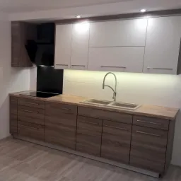 Nowoczesna kuchnia: dolne szafki z frontami w odcieniach drewna, górne szafki białe, blat imitujący drewno, czarny panel szklany nad płytą indukcyjną, oświetlenie podszafkowe LED.