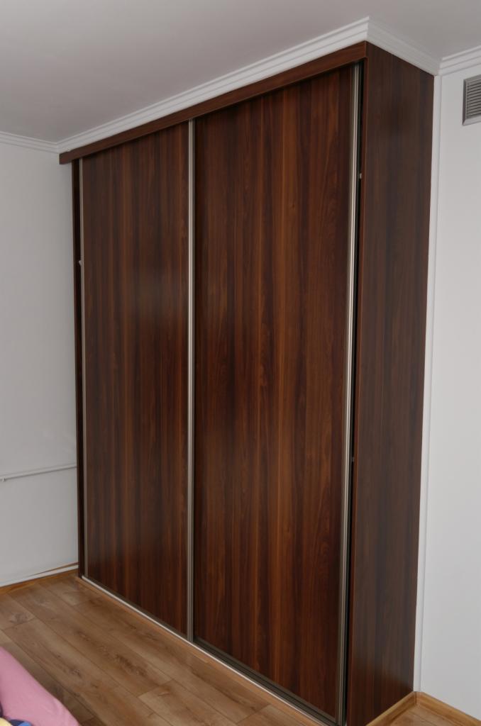 Wysoka szafa przesuwna z ciemnego drewna z metalowymi prowadnicami, dopasowana do listwy sufitowej, widoczna z perspektywy podłogi.