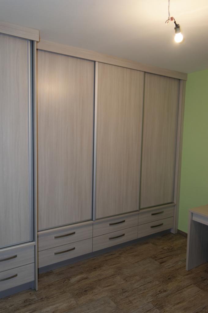 Jasnoszary regał z przesuwnymi drzwiami i trzema rzędami szuflad na tle zielonej ściany, widoczny fragment blatu biurka.