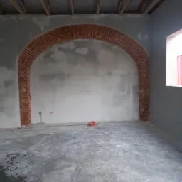 Wnętrze w trakcie prac wykończeniowych: surowe ściany z ceglanym łukiem w przejściu, betonowa podłoga, widoczne elementy drewnianej konstrukcji dachu.