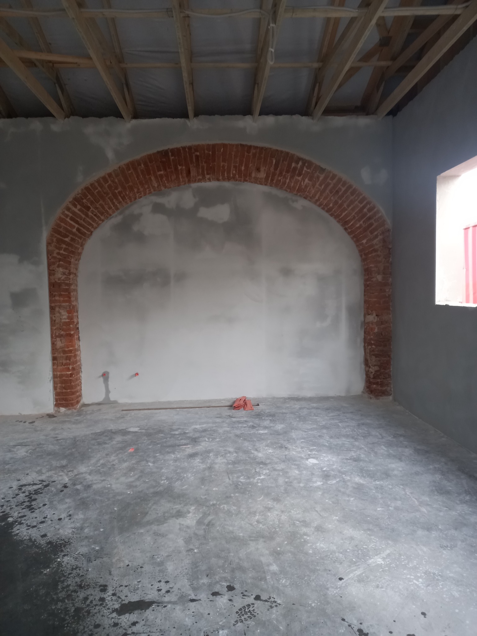 Wnętrze w trakcie prac wykończeniowych: surowe ściany z ceglanym łukiem w przejściu, betonowa podłoga, widoczne elementy drewnianej konstrukcji dachu.