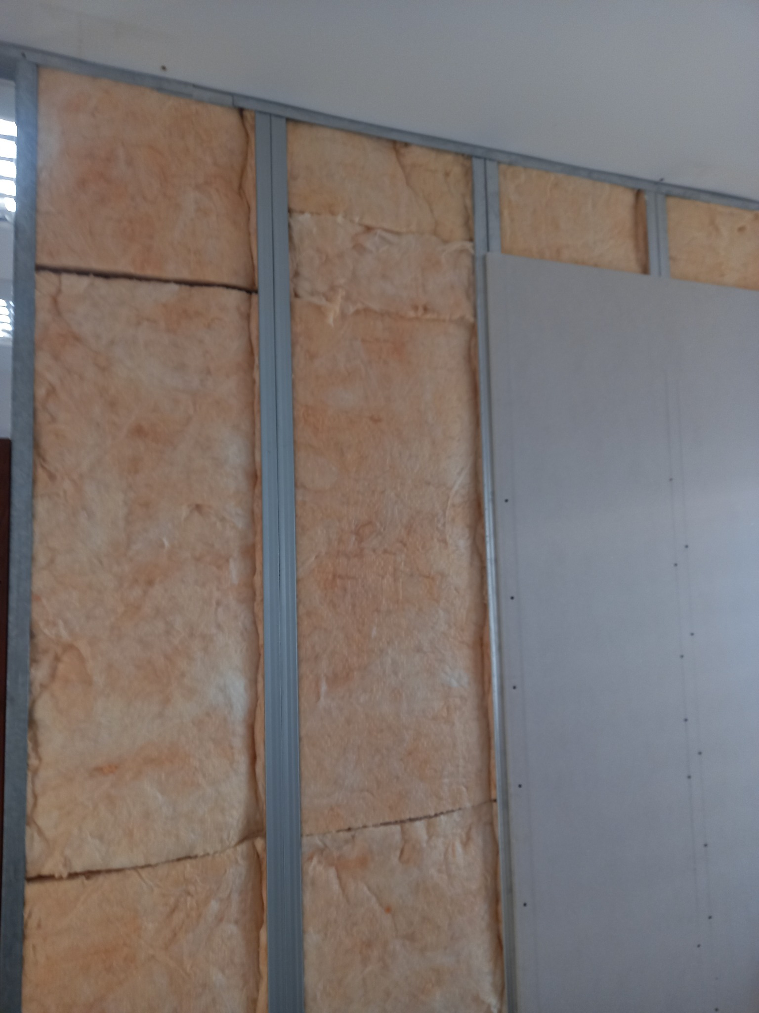 Montaż ścianki działowej z płyty karton-gips: widoczne metalowe profile, izolacja z wełny mineralnej i częściowo zamontowana płyta G-K przykręcona wkrętami.