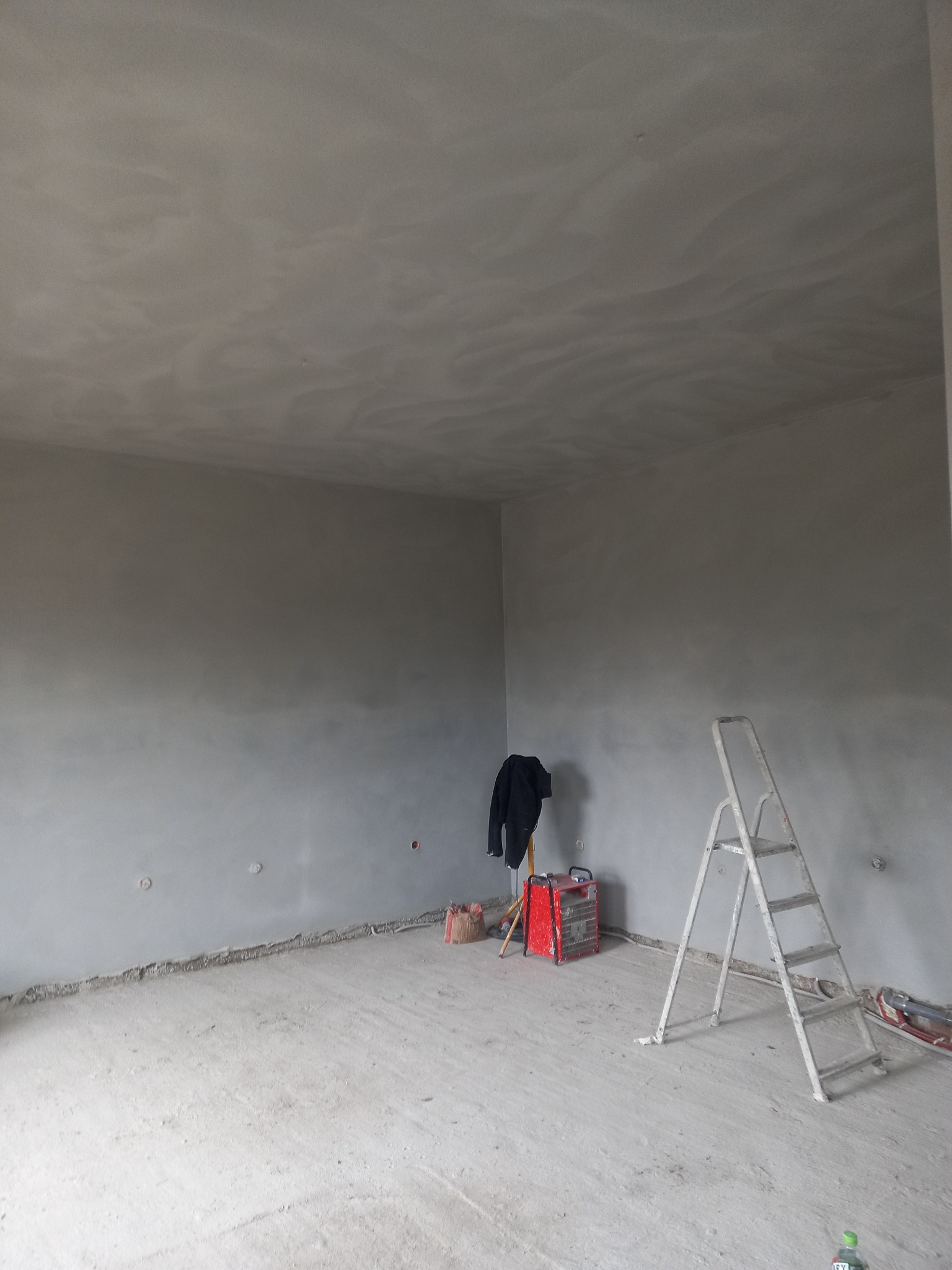 Pokój po nałożeniu gładzi, widoczne ściany i sufit z nierównomierną fakturą, drabina malarska, grzejnik elektryczny oraz ubranie robocze.