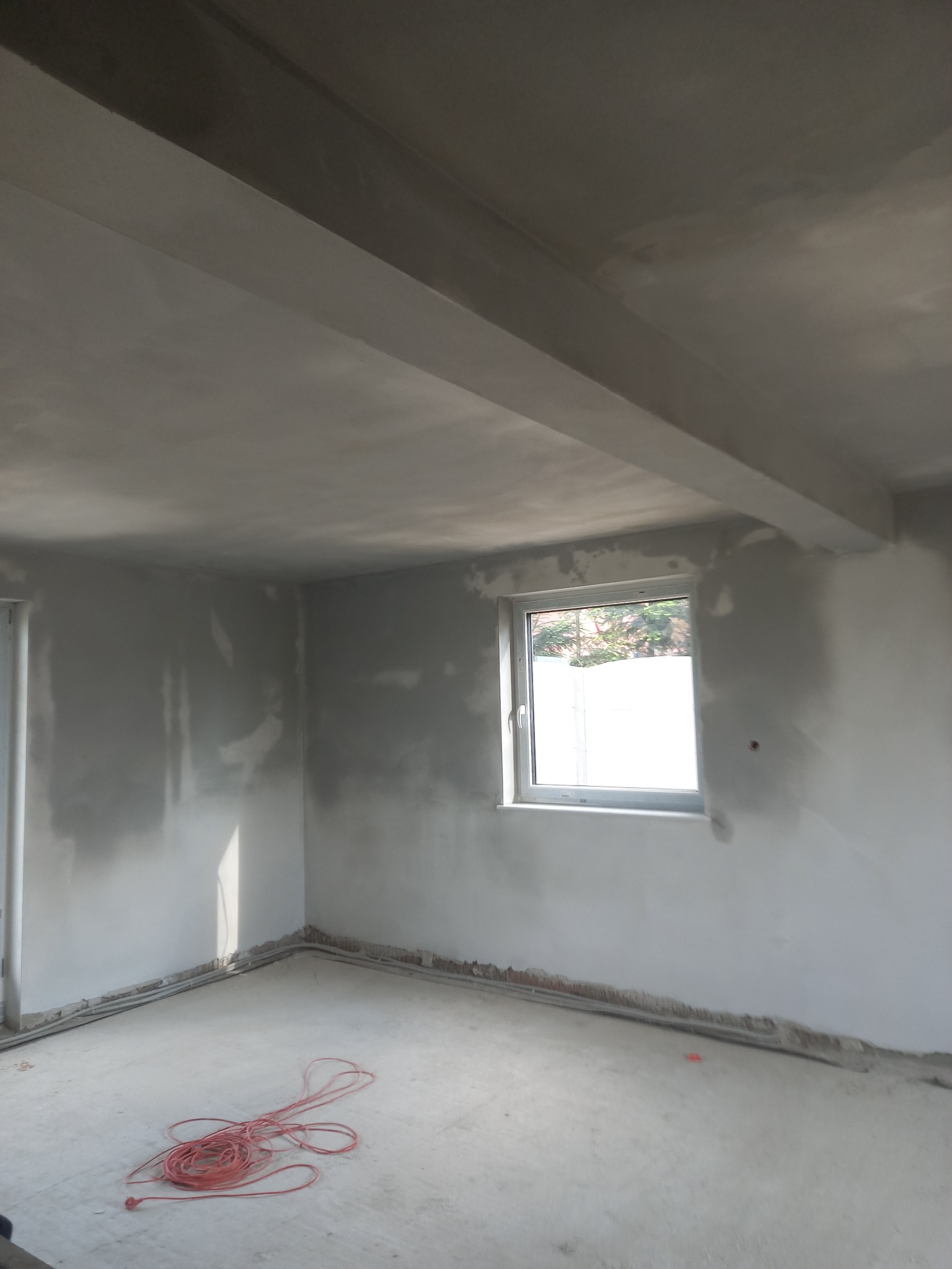Wnętrze pomieszczenia w stanie surowym po wstępnym wyrównaniu ścian i sufitu, widoczne ślady świeżych prac wykończeniowych, kabel elektryczny na podłodze.