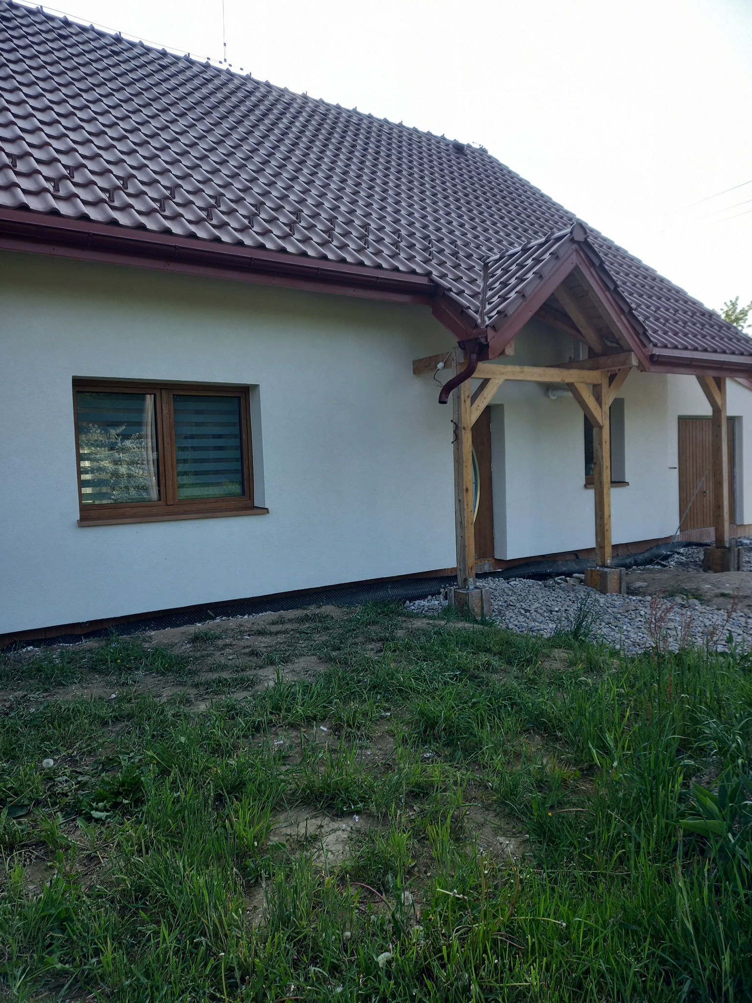 Biała elewacja domu jednorodzinnego z brązową dachówką, widoczna drewniana konstrukcja zadaszenia nad wejściem, okno z żaluzjami poziomymi i fragment trawnika.