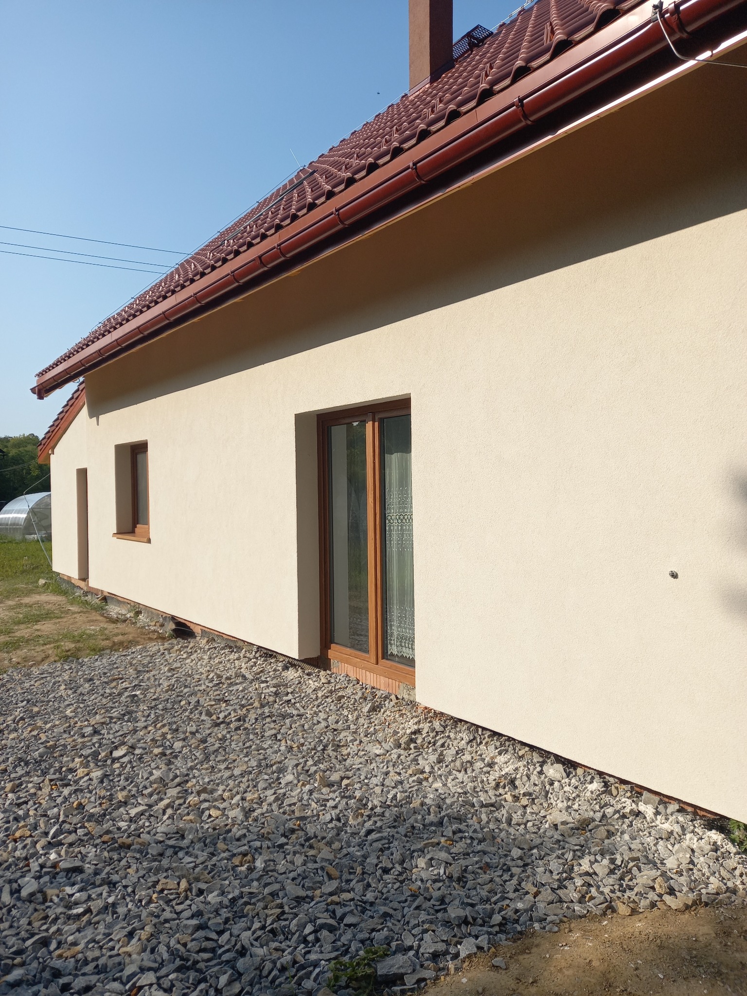 Beżowa elewacja domu jednorodzinnego z brązowymi oknami i dachem z dachówki ceramicznej, widok z boku na tle błękitnego nieba, podłoże wyłożone kamieniami.