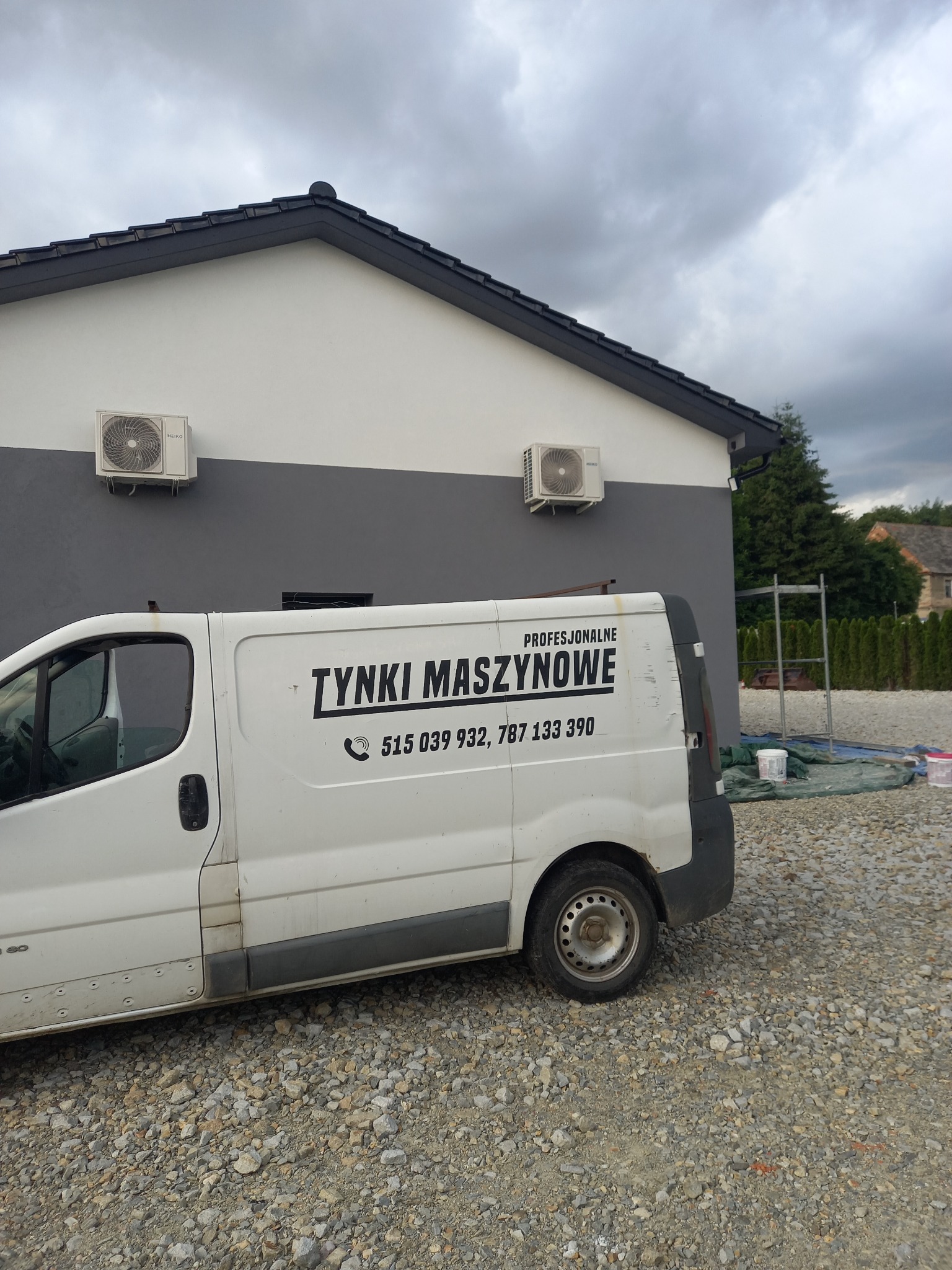 Biały samochód dostawczy z napisem 'Tynki Maszynowe' na tle dwukolorowej elewacji domu z zamontowanymi klimatyzatorami, widoczne rusztowanie i wiaderka z farbą.